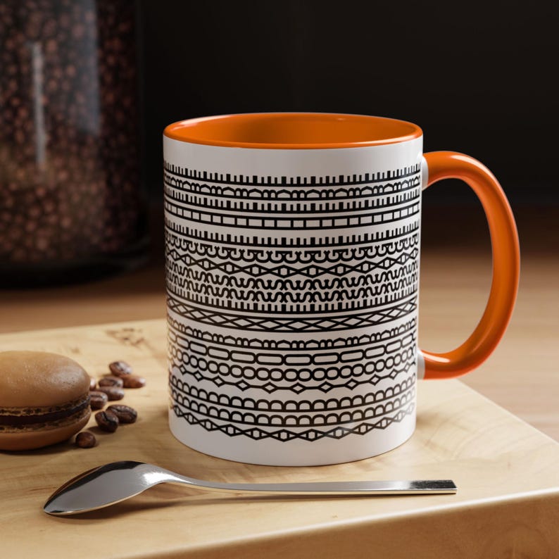 Pode incluir: Caneca de cer&acirc;mica branca com interior e al&ccedil;a laranja. A caneca apresenta um padr&atilde;o geom&eacute;trico preto no exterior. Uma colher de prata, um macaron e gr&atilde;os de caf&eacute; est&atilde;o em uma superf&iacute;cie de madeira ao lado da caneca.