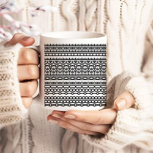 Pode incluir: Caneca de cer&acirc;mica branca com um padr&atilde;o geom&eacute;trico preto repetido. O design inclui v&aacute;rias combina&ccedil;&otilde;es de linhas e formas, criando um efeito de borda visualmente interessante. A caneca &eacute; segurada por uma pessoa que usa um su&eacute;ter creme.