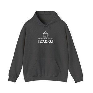 Puede incluir: Una sudadera con capucha gris oscuro que presenta un gráfico blanco de un contorno de casa relleno con patrones de circuitos impresos sobre el texto "THERE IS NO PLACE LIKE 127.0.0.1". La prenda incluye un bolsillo canguro y cordones ajustables.