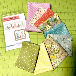 May include: A set of fabric squares in various floral patterns and colors, including pink, green, yellow, and blue. The fabric squares are arranged in a circle on a green cutting mat. A printed pattern sheet for a "Sweet Spring Bunny" runner and pillow is also included.