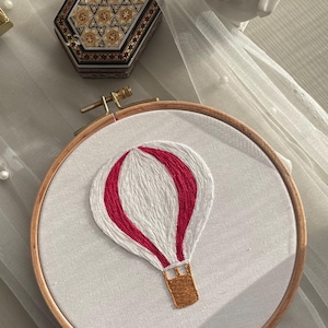 May include: An embroidery hoop with a white fabric featuring a hot air balloon design. The balloon is white with red accents, and a brown basket. A decorative box with a geometric pattern is in the background.