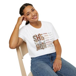 Könnte beinhalten: Weißes T-Shirt mit dem Text "SHE is MOM" in Leopardenmuster und brauner Schrift, mit floralen Akzenten. Zusätzlicher Text enthält "STRONG", "CHOSEN", "BEAUTIFUL", "CAPABLE", "VICTORIOUS", "NEVER ALONE" und "CREATED".