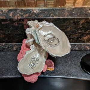 May include: A white oyster shell with a pink base, used as a decorative ring holder. The shell is open and has two rings inside. There is a small pink shell on the base.
