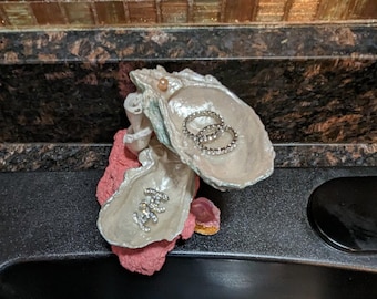 May include: A white oyster shell with a pink base, used as a decorative ring holder. The shell is open and has two rings inside. There is a small pink shell on the base.
