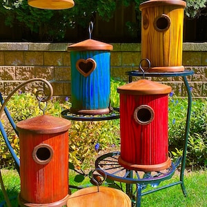 May include: Five colourful birdhouses with heart-shaped openings. The birdhouses are made of wood and are painted in different colours, including purple, blue, yellow, red, and green. They are displayed on a black metal stand.