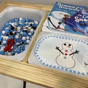 Pode incluir: Uma mesa sensorial de madeira com três compartimentos. O primeiro compartimento está cheio de itens sensoriais azuis e brancos, o segundo compartimento tem um prato branco com um desenho de boneco de neve e uma borda de floco de neve azul, e o terceiro compartimento tem uma caixa branca com pontos pretos e um pau marrom.