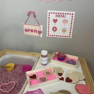 May include: A pink and white play bakery set with a sign that says "Bakery" and "Open". The set includes a menu with prices for donuts and cupcakes, a tray of cupcakes, a tray of heart-shaped cookies, a rolling pin, and a container of sprinkles.