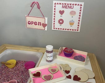 Puede incluir: Un conjunto de panadería de juguete rosa y blanco con un letrero que dice "Bakery" y "Open". El conjunto incluye un menú con precios para donas y cupcakes, una bandeja de cupcakes, una bandeja de galletas con forma de corazón, un rodillo y un recipiente de chispas.