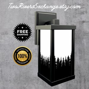 Puede incluir: Lámpara de pared exterior negra con un diseño rectangular. Cuenta con un marco negro y un panel blanco con una silueta de bosque negro. El nombre de la tienda Etsy "Two Rivers Exchange" está en la parte superior.