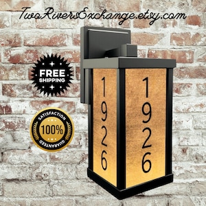 Puede incluir: Luminaria de pared exterior negra con un panel rectangular dorado que muestra el número "1926" en negro. La luminaria está montada en una pared de ladrillo. El nombre de la tienda Etsy "Two Rivers Exchange" está en la parte superior.