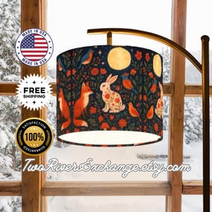 May include: A floor lamp with a cylindrical lampshade featuring a whimsical forest design. The lampshade showcases foxes, rabbits, birds, flowers, and moons against a dark blue background. The lamp has a gold-toned arched arm and a wooden base.