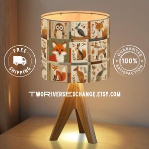 May include: A wooden table lamp with a lampshade displaying a patchwork design of woodland creatures, such as foxes, owls, and rabbits. The lamp has a three-legged wooden base. The Etsy shop name is visible on the base.