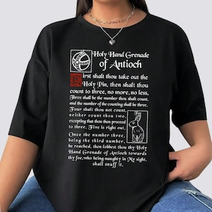 May include: A black short-sleeved t-shirt featuring a white graphic print of the Holy Hand Grenade of Antioch text and a rabbit illustration. The text includes instructions for counting to three and mentions the foe, all in a classic font style.