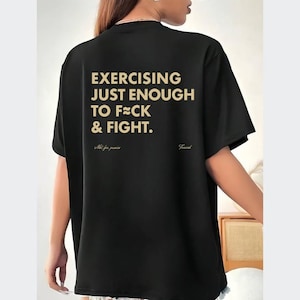 May include: Black t-shirt with gold text that reads "EXERCISING JUST ENOUGH TO F**K & FIGHT." Below the text are the words "Not for primas" and "Found". The shirt is a casual style.