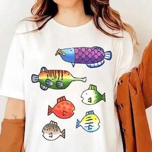 May include: White t-shirt featuring colorful cartoon fish illustrations. The fish designs include a purple and blue fish, a green and orange fish, and several smaller fish in red, green, yellow, and brown.