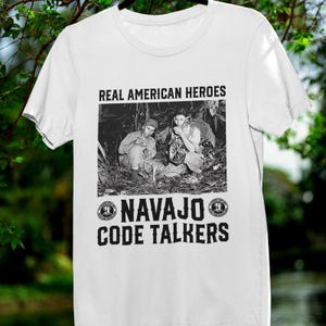 May include: White t-shirt featuring a black and white image of two people in a forest setting. The text "REAL AMERICAN HEROES" is above the image, and "NAVAJO CODE TALKERS" is below it in a bold font.