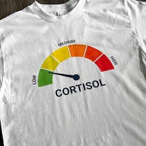 May include: White t-shirt with a graphic of a cortisol meter. The meter has a needle pointing to the "LOW" section, with a color gradient from green to red, and the word "CORTISOL" below the meter.