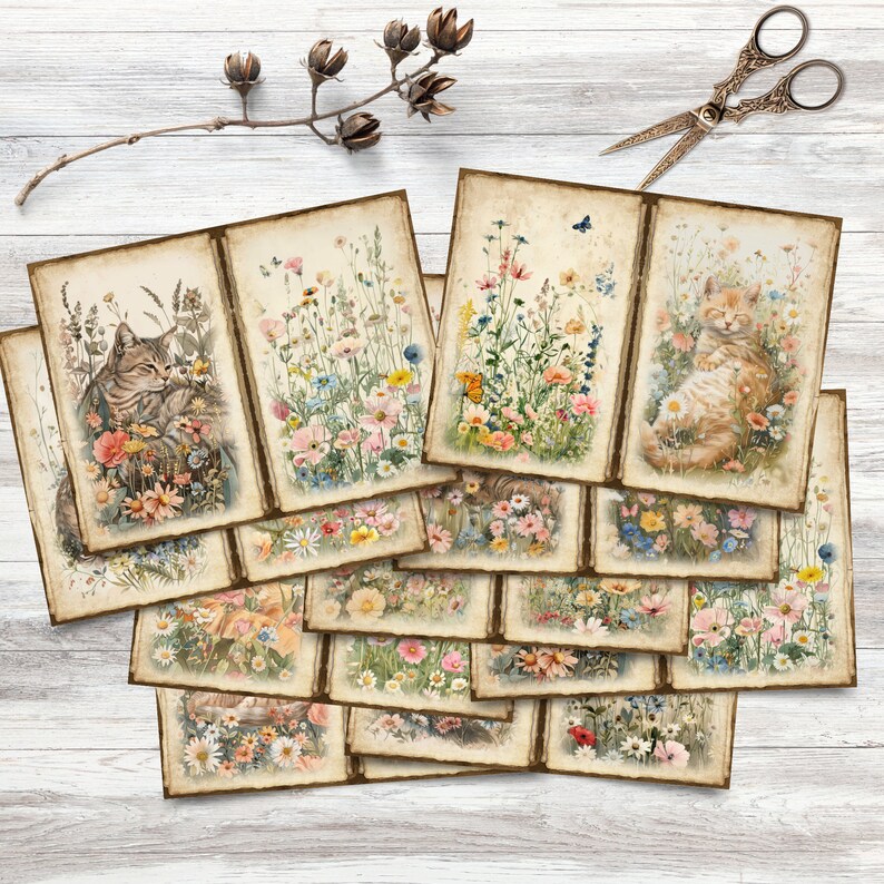 May include: A collection of vintage-style cards featuring watercolor illustrations of cats in flower fields. The cards showcase a variety of colorful wildflowers and butterflies, with a distressed, aged paper effect. A pair of decorative scissors and dried flowers are also visible.
