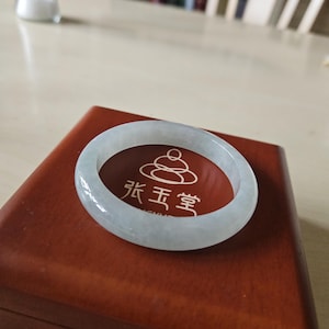 Puede incluir: Un brazalete de jade blanco pálido, liso y translúcido, sobre una caja de madera marrón oscuro pulida. La caja presenta una incrustación circular roja con un gráfico blanco de piedras apiladas y los caracteres chinos 张玉堂 en blanco.
