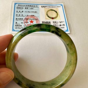 Könnte beinhalten: Ein glatter, kreisförmiger Jade-Armreif mit einem gesprenkelten Muster aus tiefem Waldgrün und durchscheinenden hellgrünen Farbtönen. Das Schmuckstück wird von einem weißen Identifikationszertifikat mit blauem Rand begleitet, das 74,64 g angibt.
