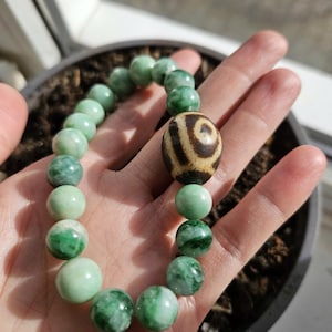 May include: A beaded bracelet featuring light green and dark green jade beads. The bracelet includes a large, patterned bead with brown and cream colors. The bracelet is held in a hand.