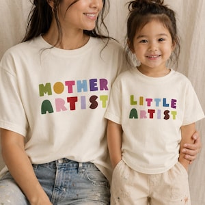 以下が含まれることがあります： クリーム色の半袖Tシャツ2枚。大きい方のTシャツには、カラフルで丸みを帯びたブロック体で「MOTHER ARTIST」とプリントされています。小さい方のTシャツには、同じスタイルのカラフルなブロック体で「LITTLE ARTIST」とプリントされています。
