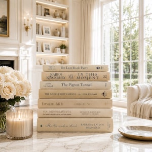 May include: A stack of seven hardcover books with cream-coloured spines, featuring titles including The Last Book Party, In This Moment, The Pigeon Tunnel, The Book of Lost Friends, Prodigal Son, You: Being Beautiful, and The Cavendon Luck, arranged on a table.