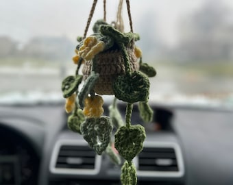Op de afbeelding: Een handgemaakte gehaakte hangplant met een bruine pot, gele bloemen en groene bladeren. De plant wordt opgehangen met een koord van natuurlijke vezels. De bladeren zijn in verschillende groentinten en de bloemen zijn geel.