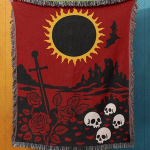 May include: Red throw blanket with a black and yellow sun, castle, sword, roses, bat, and skull design. The blanket has a fringed edge and is held up against a colorful wall. Digital Cottage Crafts logo is in the top left corner.