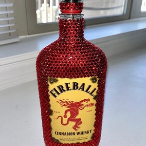 May include: A red glass bottle covered in red rhinestones with a yellow label that reads "FIREBALL", "RED", "HOT", "CINNAMON WHISKY", "ALC. 33% BY VOL. (166 PROOF)", and "WHISKY WITH NATURAL CINNAMON FLAVOR".