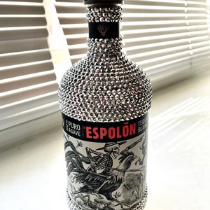 May include: A silver bottle of Espolón Tequila Blanco, covered in rhinestones, with a black and white label featuring a skeleton riding a rooster.