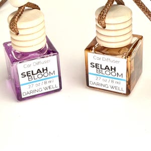 May include: Two car diffusers with wooden tops and brown cords. One is purple, the other is amber. Each has a label that reads "SELAH BLOOM" and "DARING WELL". The bottles are square and contain 8 ml of liquid.