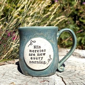 May include: A ceramic mug with a blue-green glaze and a speckled white circular center panel. The text "His mercies are new every morning." is printed in black on the panel, accompanied by small decorative flourishes at the top and bottom.
