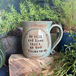 Puede incluir: Una taza de cerámica turquesa con asa, con la inscripción "Be still and know that I am God Psalms 46:10" dentro de una corona. La taza está sobre un bloque de madera, rodeada de plantas verdes.