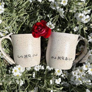 May include: Two speckled off-white ceramic mugs with handles, one featuring the text MR and the other featuring the text MRS in black lettering. A single vibrant red rose is positioned between the two mugs, resting on the ceramic surfaces.