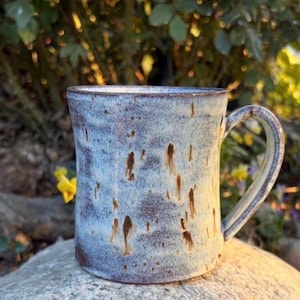 May include: A handcrafted ceramic mug with a speckled blue and brown glaze. The mug has a rounded shape and a sturdy handle. The exterior features a mottled pattern, and the interior is a lighter shade of blue. Ideal for tea or coffee.