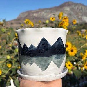 May include: A white ceramic planter with a ruffled rim and a matching saucer. The exterior features a hand-painted dark blue mountain range design with small white speckles resembling stars, contrasted against a lighter gray mountain reflection.