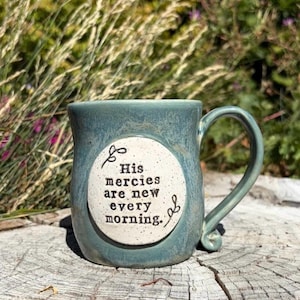 May include: A ceramic mug with a glossy blue-green glaze and a speckled off-white circular medallion on the front. The medallion features the black printed text, His mercies are new every morning, with decorative flourishes above and below the words.