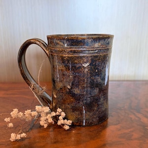 May include: A handcrafted ceramic mug with a dark brown, glossy glaze. The mug features a sturdy handle and a textured surface, with subtle variations in color. A small sprig of dried flowers rests near the mug.