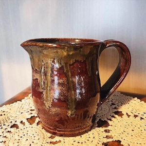 May include: A ceramic pitcher with a glossy finish, featuring a deep reddish-brown base color and a contrasting metallic gold glaze that drips from the rim down the sides. The vessel includes a curved handle and a small pouring spout at the top.