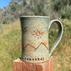 Puede incluir: Taza de cerámica hecha a mano con un esmalte verde claro y un diseño moteado marrón. La taza presenta un diseño de montaña y sol, con la palabra "tonasket" en la base. La taza está sobre un poste de madera.