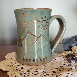 以下が含まれることがあります： 薄い青色のセラミック製のマグカップで、茶色の山の風景と太陽のデザインが施されています。マグカップの底には「tonasket」という文字が書かれています。