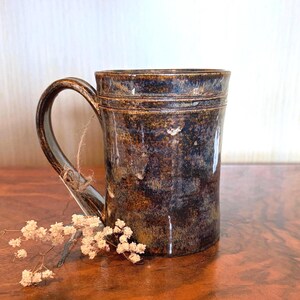 May include: A cylindrical ceramic mug with a dark, mottled glaze featuring deep brown, black, and subtle blue tones. The mug has a curved handle with a small piece of twine tied around it and a horizontal grooved line near the top rim.