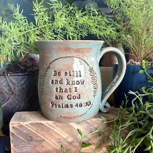 May include: A ceramic mug with a light blue and brown glazed finish featuring a circular embossed design with the text Be still and know that I am God Psalms 46:10. The mug has a curved handle and a smooth, rounded rim with a glossy texture.
