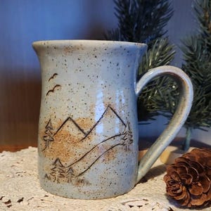 May include: A handmade ceramic mug with a light blue speckled glaze. The mug features a mountain and tree design in brown and black, with three birds flying near the rim. The handle is curved, and the mug is suitable for hot or cold beverages.
