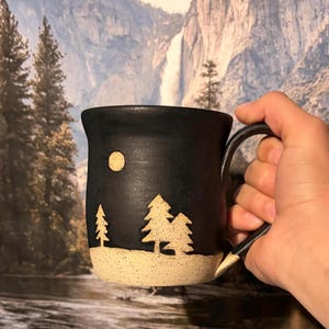 May include: A black ceramic mug featuring a carved design of three evergreen trees and a circular moon against a dark sky. The base of the mug is a speckled cream color, contrasting with the matte black upper portion and handle.