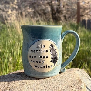 Puede incluir: Una taza de cerámica turquesa con asa curva. La taza presenta el texto "His mercies are new every morning" en un óvalo color crema, junto con una ilustración de pluma negra. La taza está sobre una roca.