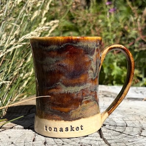 May include: A ceramic mug with a glossy glaze featuring swirling patterns of deep brown, amber, and muted green. The base is a contrasting matte cream colour with the word "tonasket" stamped in lowercase black letters near the bottom rim.