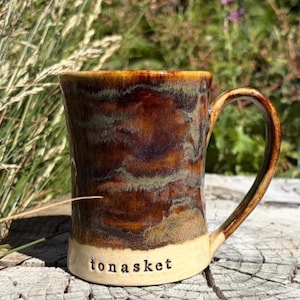 May include: A ceramic mug featuring a glossy, multi-toned glaze in shades of deep brown, amber, and muted green. The base of the mug is a smooth, unglazed cream color with the word "tonasket" stamped in lowercase black letters on the front.