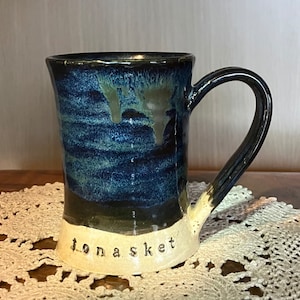 Könnte beinhalten: Eine blaue und weiße Keramiktasse mit Henkel. Die Tasse hat eine dunkelblaue Glasur mit grünen und weißen Streifen. Der Boden der Tasse ist weiß mit dem Wort "jonaske" in Schwarz geschrieben.